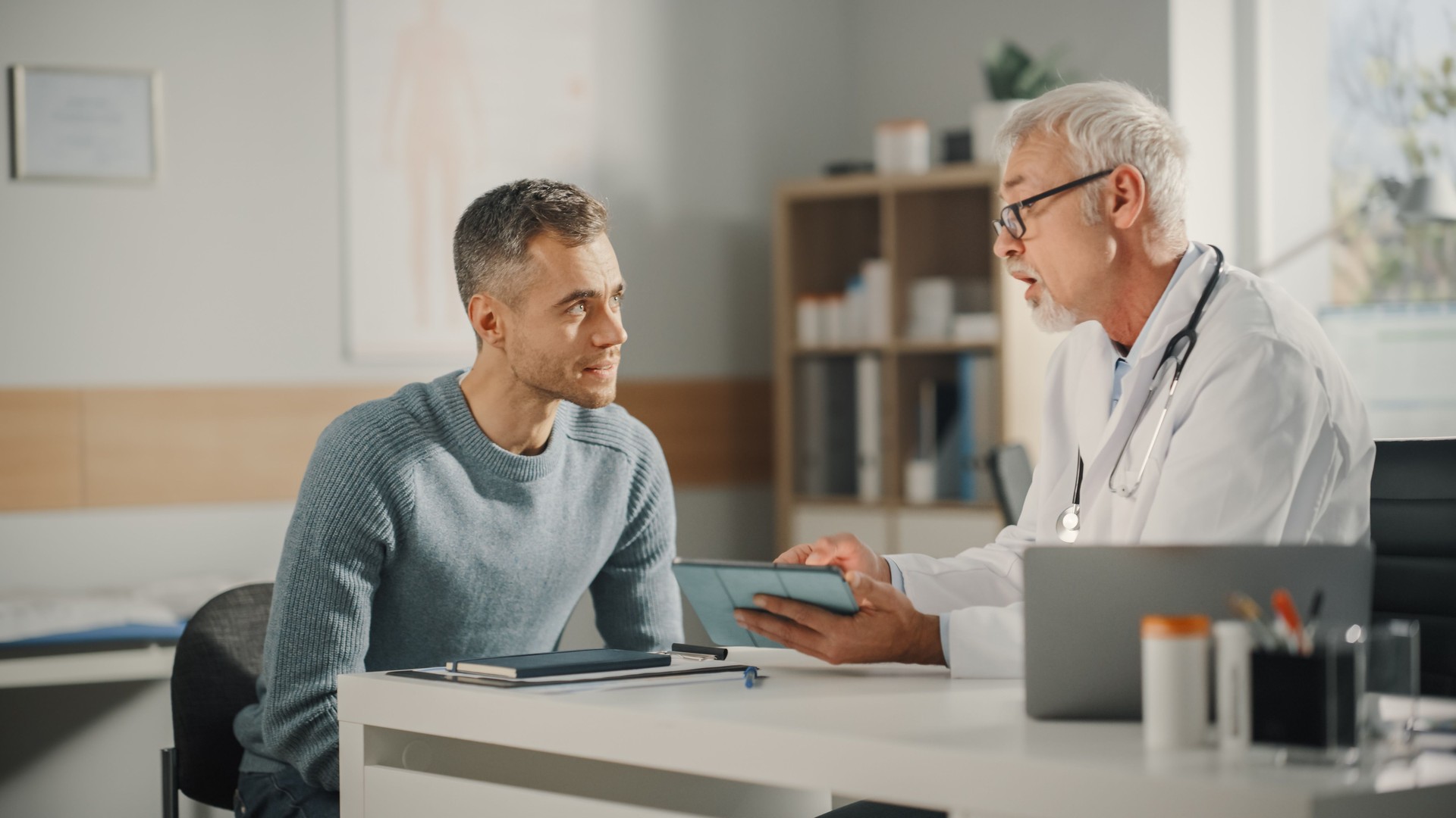 Experienced Middle Aged Family Doctor Showing Analysis Results on Tablet Computer to Male Patient During Consultation in a Health Clinic. Physician Sitting Behind a Desk in Hospital Office.